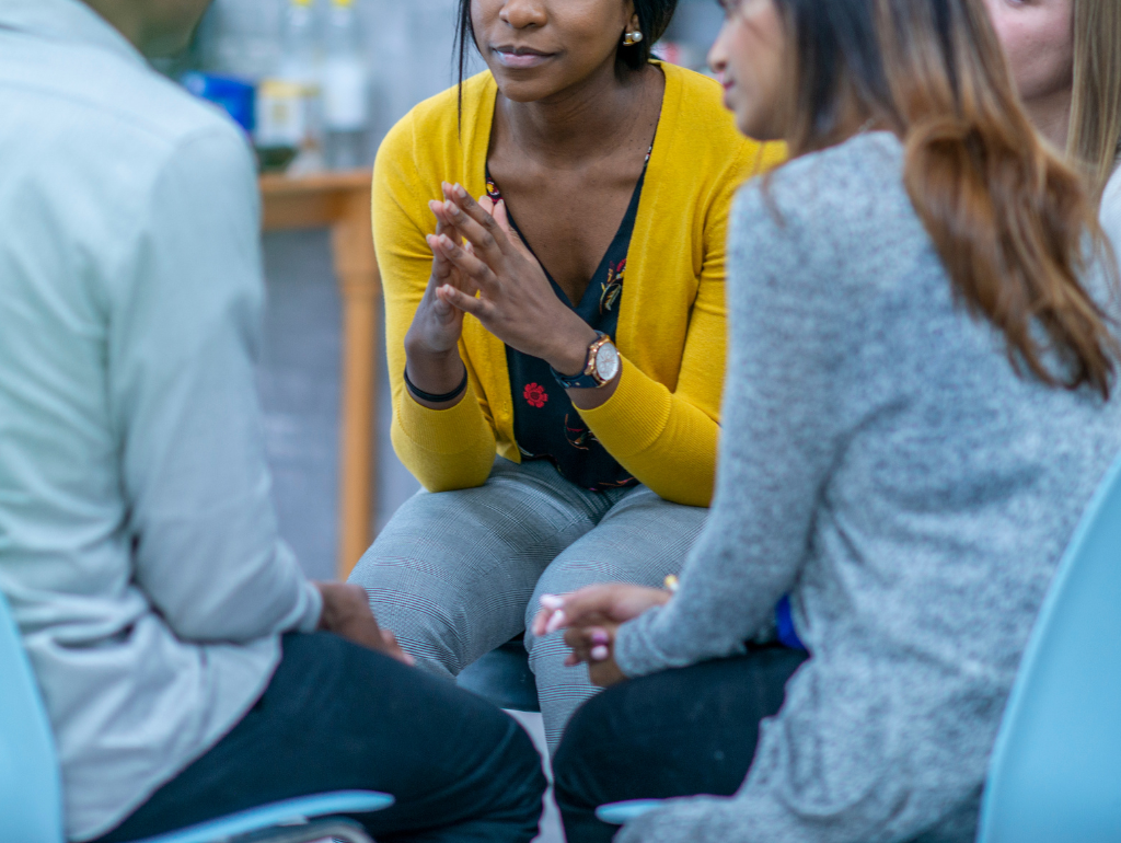 A group of people sitting together, someone has their hands together in a listening pose