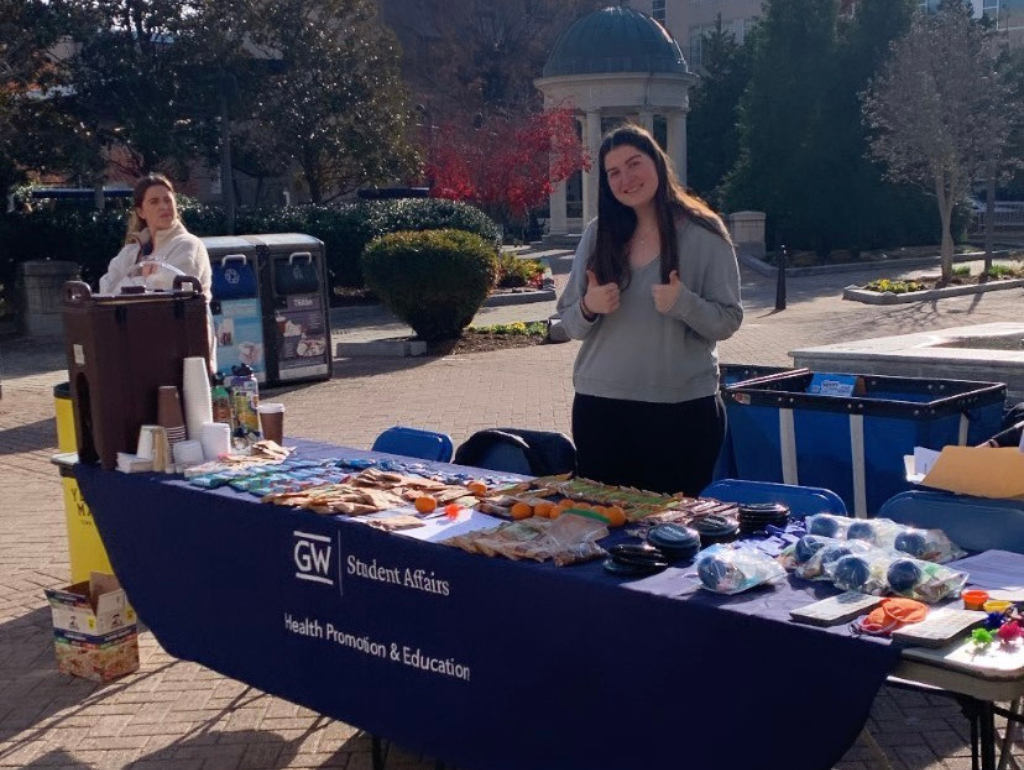 A student smiling and giving "two thumbs up" while volunteering at a Health Promotion table in Kogan Plaza