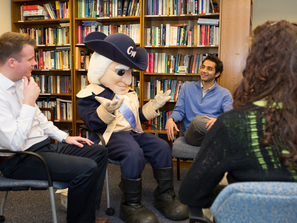 George mascot sitting in a circle with other students; his arms are shrugging